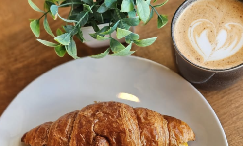 A steak-and-egg breakfast croissant with melted cheese and caramelized onions on a white plate, beside a latte with heart-shaped foam art.
