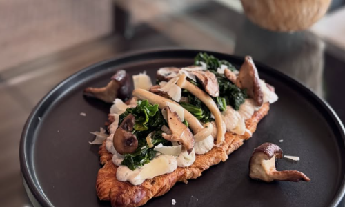 An open-face croissant topped with sautéed kale, wild mushrooms, and shaved parmesan on a dark plate.